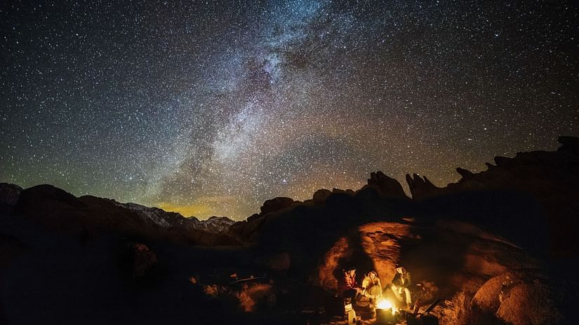 A group of people huddled next to a bonfire with the milky way seen in the background.