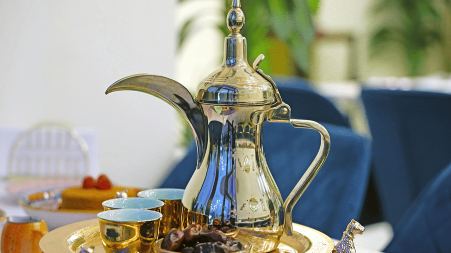 A view of a stainless steel Dallah Coffee Pot, one of the best souvenirs from Qatar to buy, kept on a table with small glasses next to it.