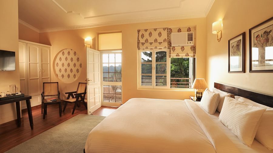 An image of a Super Deluxe Room featuring a double size bed, two chairs, a table where coffee maker is kept, and an open door from where sunlight is flooding the room - Lamrin Boutique Cottages Rishikesh