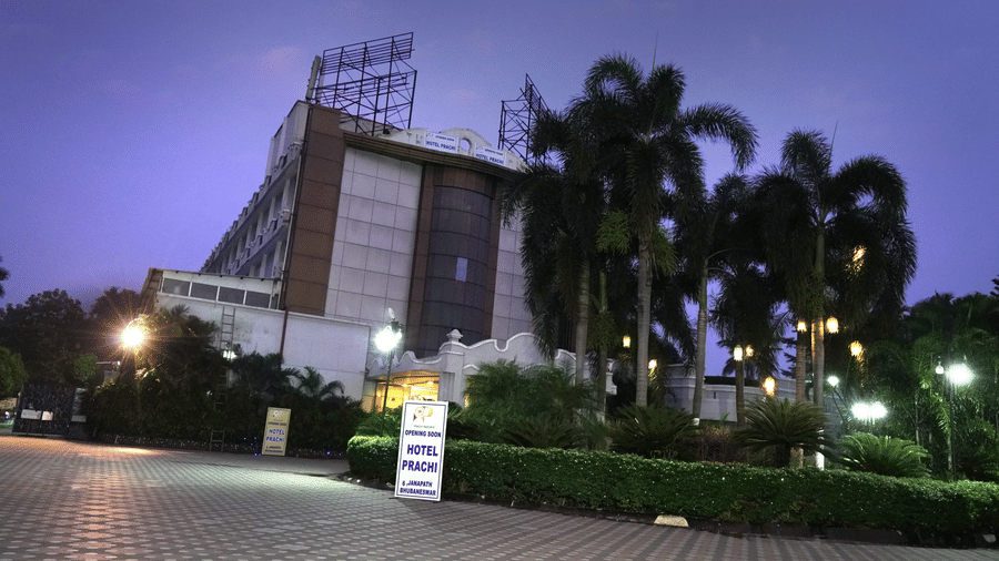 Facade of Sarovar Portico, Bhubaneswar, seen under the dark sky, with a well-lit driveway, a manicured garden, and a board with the name of the hotel on it.
