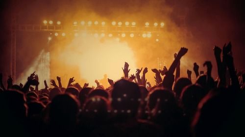 A crowd of people at a concert with their hands raised, against a brightly lit stage.