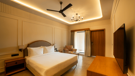A neat room featuring a king size bed with a brown headboard, a wooden TV console, a ceiling fan, and floor-to-ceiling window at Jivana Wellness Retreat, Bhopal.