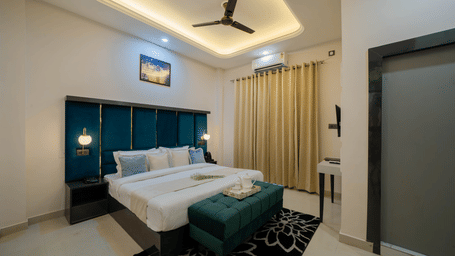 A bedroom with a king size bed, bedside tables, curtains, ceiling fan, wall art, and a bench at Perfectstayz Value Aalaya Suites, Haridwar.
