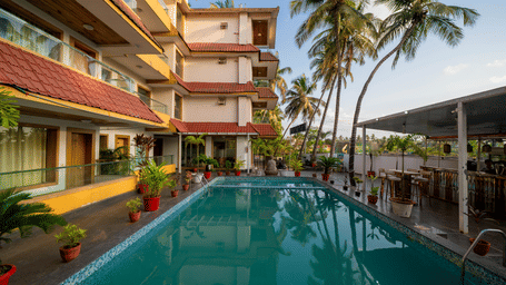 A sunny outdoor swimming pool is located within the courtyard surrounded by palm trees and buildings at Perfectstayz Premium Goa Continental.