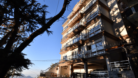 A facade of Perfectstayz Value Shimla (Namah Retreat) with balconies viewed from below against a clear sky and a tree in foreground.