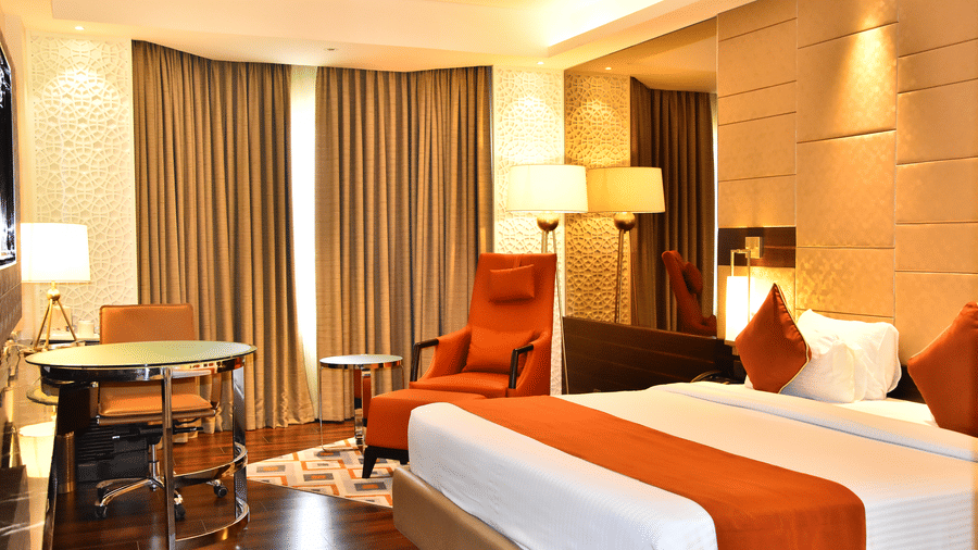 A spacious room with a large bed, a cushioned chair with a table beside it, and windows covered with curtains | Cenneys Gateway - Pride of Salem.