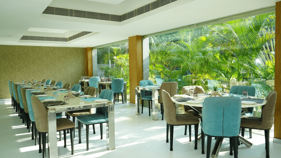 Indoor dining area with elegant table arrangements and large windows showcasing green coconut trees - Grande Bay Resort & Spa, Mamallapuram