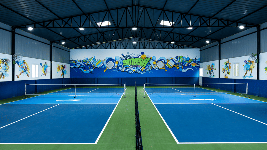 Symmetrical view of multiple indoor pickleball courts with blue and green flooring and colourful wall art, at Jenneys Residency, Coimbatore.
