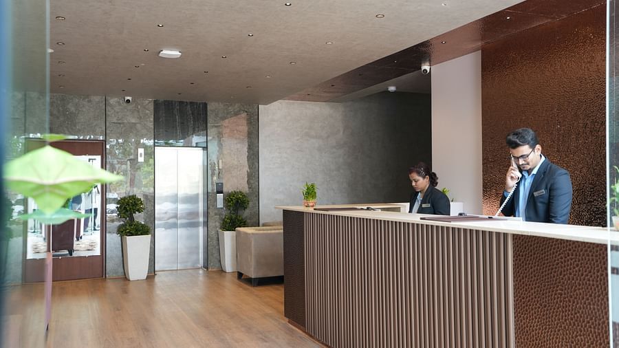 Reception area with two staff members behind the counter and a lift in the background at TGI Livancia Hillside, JC Nagar, Mysore