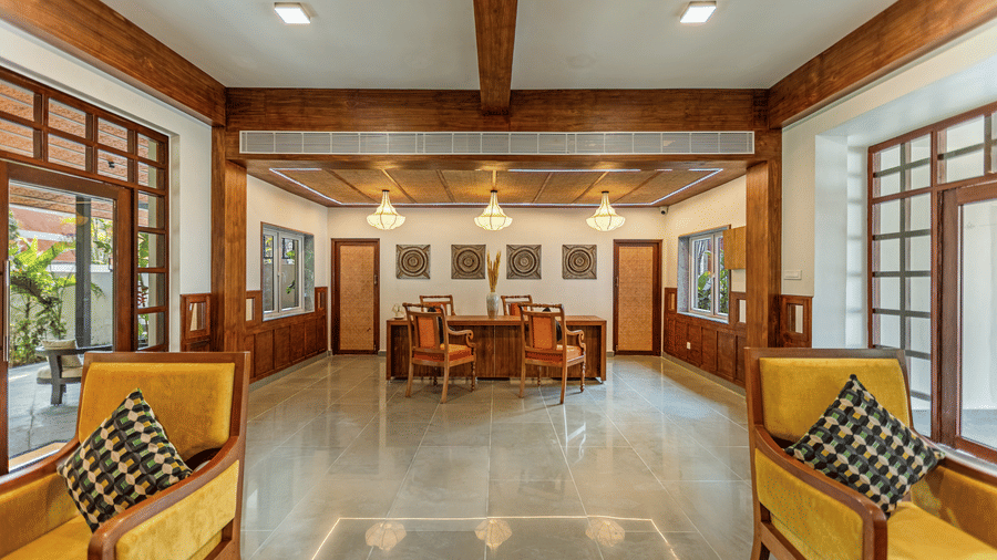 A well-lit hotel reception area with exposed wooden beams, grey tiled floors, yellow armchairs with patterned pillows, and a reception desk with orange chairs - Mango Hill Resort, Coimbatore