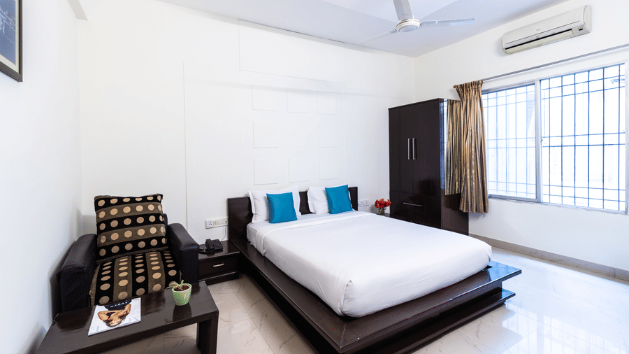 A room with a bed, an armchair, a wardrobe, and a large window at Sanctum Suites Domlur, Bangalore.