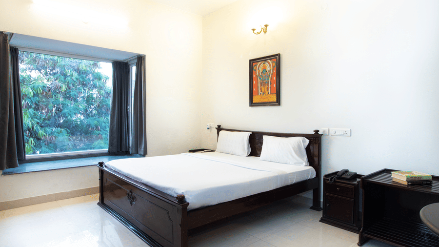 Hotel room with a double bed, traditional wall art, and a window overlooking a garden at Sanctum Suites Indiranagar, Bangalore.