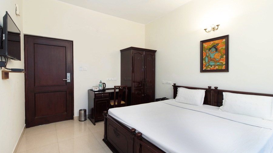 Spacious hotel room with a double bed, a dark wooden wardrobe, and traditional wall art at Sanctum Suites Indiranagar, Bangalore.