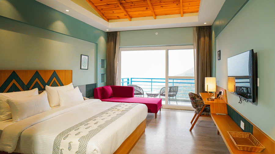 A spacious hotel room includes a large bed with white bedding, a wooden headboard, and a purple chaise lounge at the foot, with a balcony visible through large glass doors | Deluxe Room with Valley View | Sukama Resort & Spa, Muktheswar