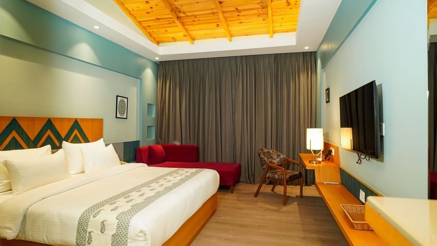 A hotel room with a large bed featuring white linens and a wooden headboard also includes a wall-mounted television and a view of curtains and a patterned wall | Deluxe Room with Valley View | Sukama Resort & Spa, Nainital