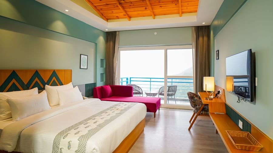 A spacious hotel room includes a large bed with white bedding, a wooden headboard, and a purple chaise lounge at the foot, with a balcony visible through large glass doors | Deluxe Room with Valley View | Sukama Resort & Spa, Nainital