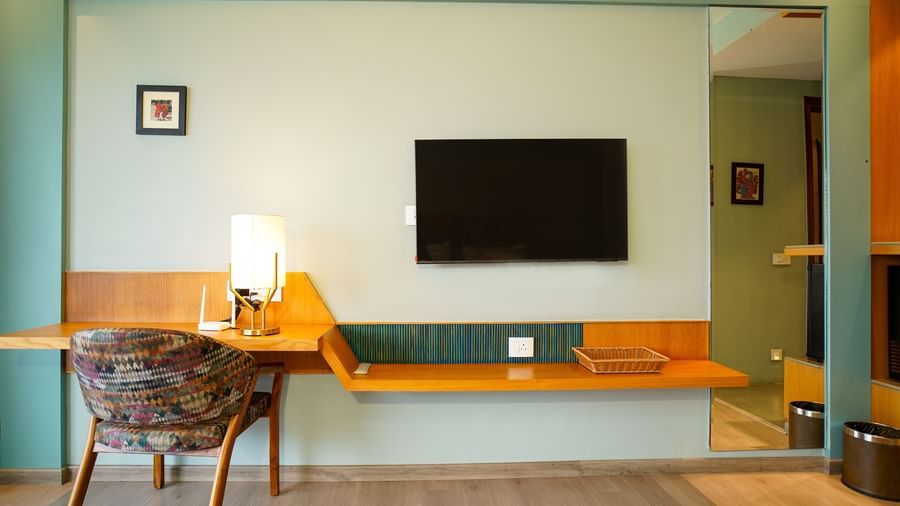 A hotel room showcases a large wall-mounted television above a long desk with a chair and lamp, complementing a bed with a wooden headboard and a white duvet | Deluxe Room with Valley View | Sukama Resort & Spa, Nainital