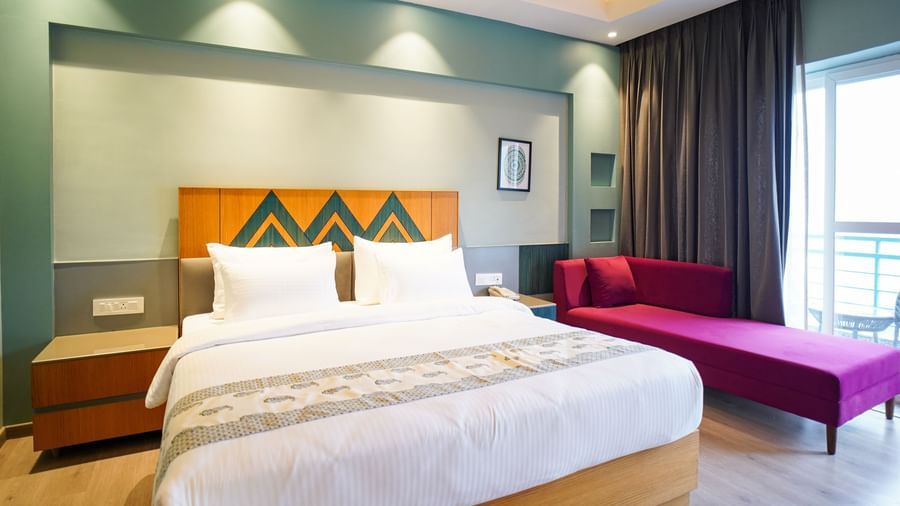 A hotel room displays a neatly made bed with white linens and a wooden headboard, accompanied by a purple chaise lounge and floor-to-ceiling curtains | Deluxe Room with Valley View | Sukama Resort & Spa, Nainital