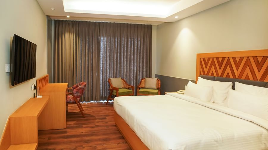 A modern hotel room with a large bed, wooden headboard, and seating area, lit by warm overhead lighting | Deluxe Room | Sukama Resort & Spa, Nainital