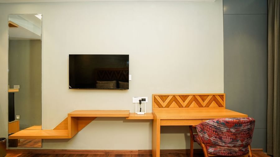 A hotel room featuring a wall-mounted television, a long wooden desk with a chair, and a patterned headboard | Deluxe Room | Sukama Resort & Spa, Nainital