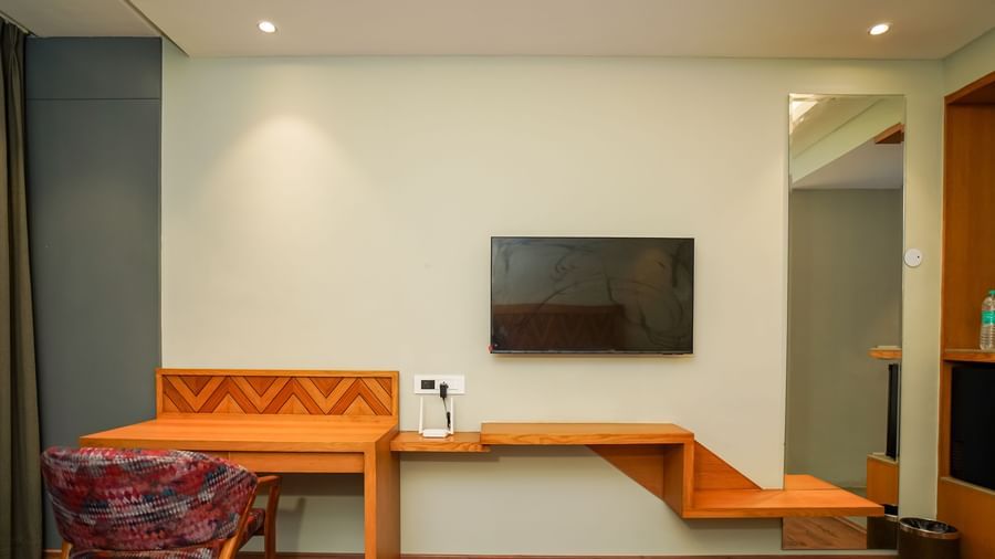 A modern hotel room with a long wooden desk and shelving unit, a chair, and a wall-mounted television | Deluxe Room | Sukama Resort & Spa, Nainital