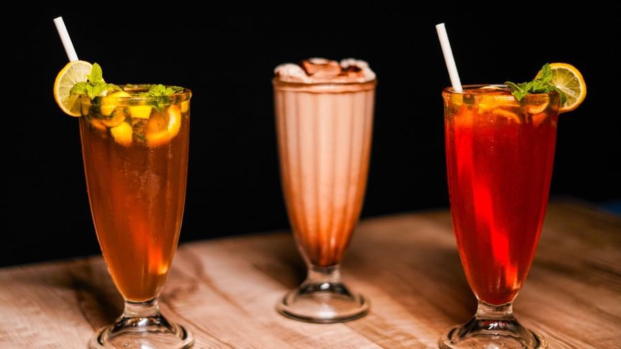 Three tall glasses containing dark beverages, each garnished with a lime slice and a straw, on a wooden surface with a dark background | Sukama Resort & Spa, Nainital
