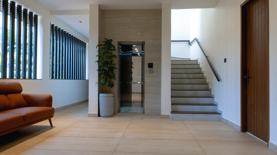 A hallway with tile flooring, a leather sofa, a tall potted plant, an elevator door, and a staircase at SUKAVI, offering access to different areas of the property.