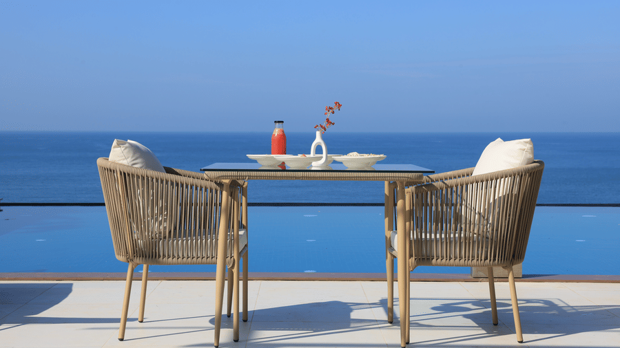 An outdoor dining table and 2 woven chairs set on a patio next to a pool, looking out over an ocean, at SUKAVI.