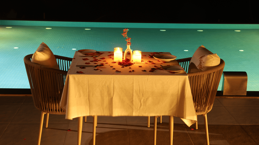 A dining table with 2 chairs, and lit candles at night, situated next to a illuminated swimming pool at SUKAVI.