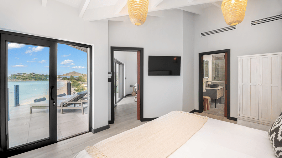 A bedroom with a king size bed, lamps, a wardrobe, a television, and glass doors leading to a terrace with an ocean view at Tamarind Hills Resort and Villas, Antigua.
