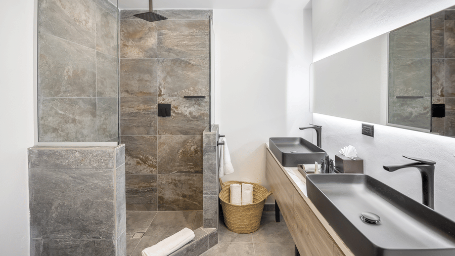A bathroom at Tamarind Hills Resort and Villas, Antigua, featuring a stone tiled shower area, toiletries, a towel, dual sinks, and a large mirror.