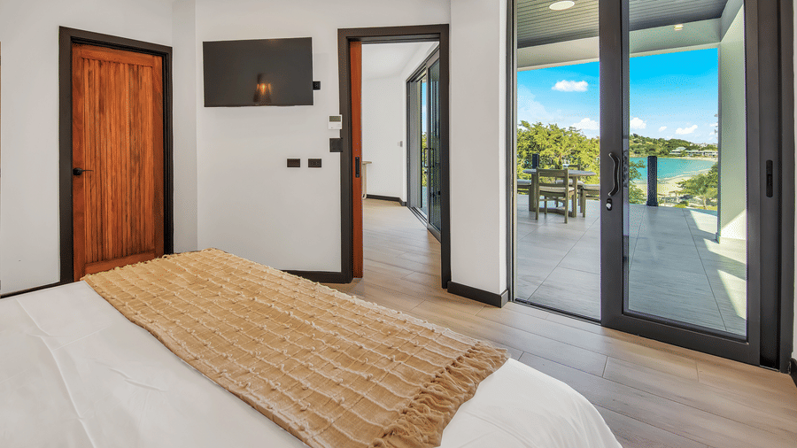 A bedroom at Tamarind Hills Resort and Villas, Antigua, with a bed, a wall mounted television, a glass sliding door leading to a balcony with a table and chairs.
