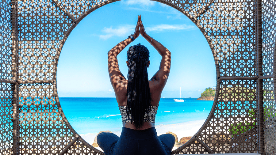 A woman on a daybed at Tamarind Hills Resort and Villas, Antigua, seated with arms lifted in a praying pose toward the sea.