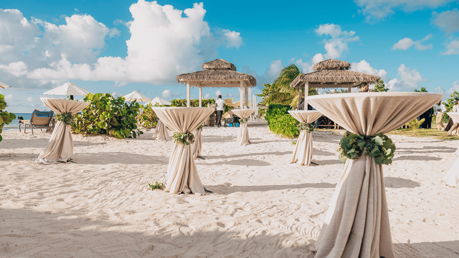 A beach setup with tall tables covered in cloth and decorated with greenery, with canopies and umbrellas in the background at Tamarind Hills Resort and Villas, Antigua.
