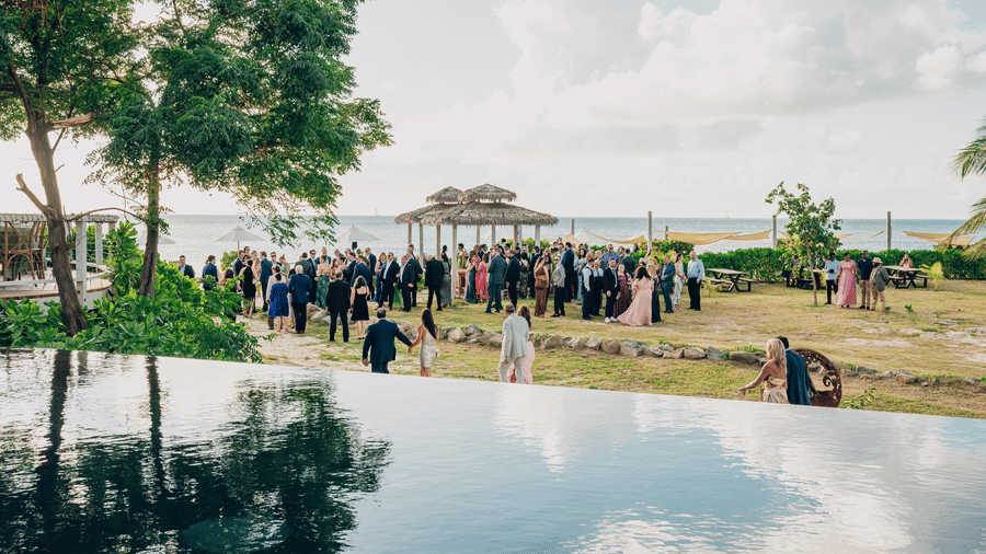 An event at Tamarind Hills Resort and Villas, Antigua with guests gathered on a lawn near a wooden gazebo overlooking the ocean, framed by lush trees and an infinity pool.