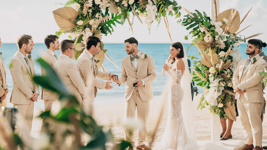 A wedding ceremony with a couple standing under an arch decorated with flowers and leaves and guests standing nearby at Tamarind Hills Resort and Villas, Antigua.