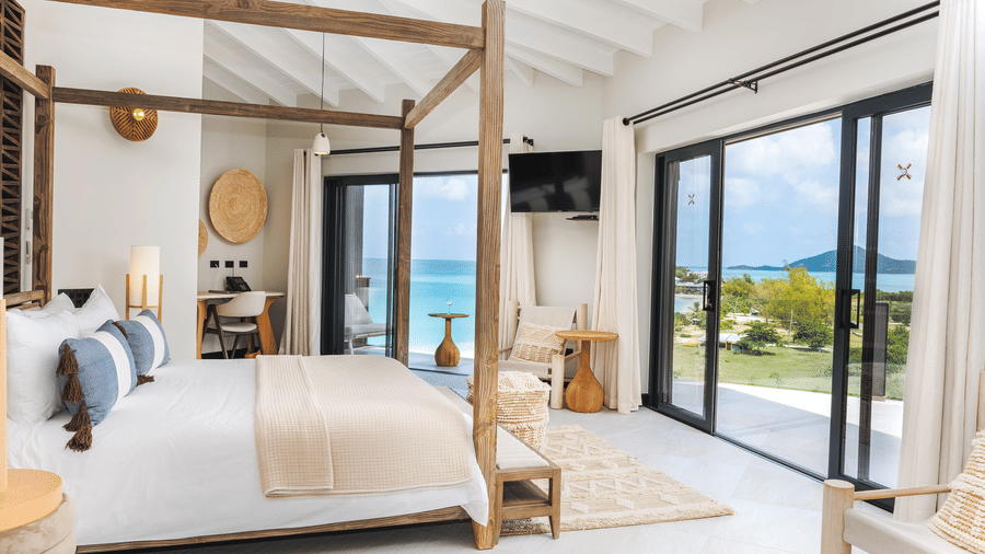 A bedroom with a four-poster bed, chairs, a desk, a television, and glass doors opening to a view of the sea at Tamarind Hills Resort and Villas, Antigua