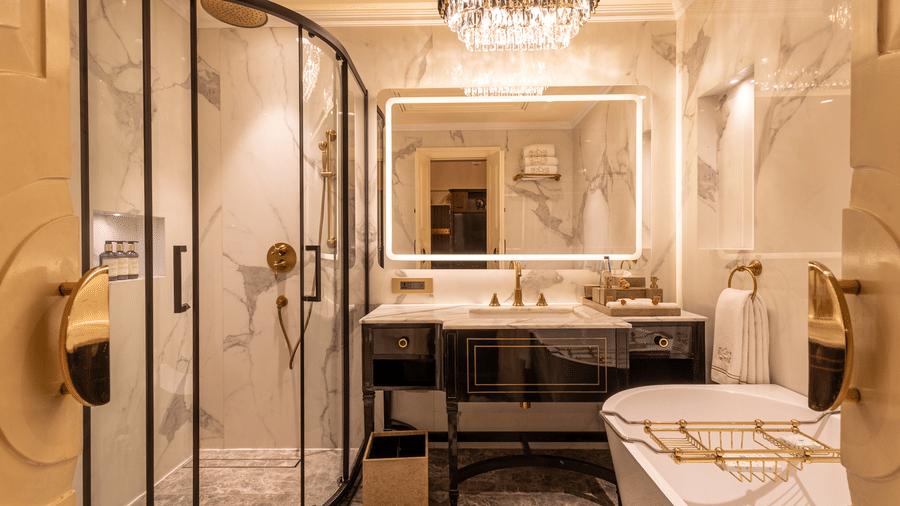 Executive Suite washroom at The Claridges New Delhi, featuring a sleek sink, wall-mounted mirror, glass-enclosed bath area, and a chandelier
