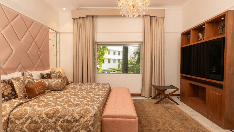 Luxury Suite at The Claridges New Delhi, featuring a cosy bed, wall-mounted TV, chandelier overhead, and a serene view through the window