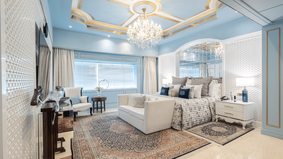 An image of a suite with a large bed and a sofa placed in front of a television and a chandelier hanging from the ceiling