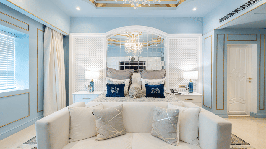 An image of a couch with cushions placed in front of a bed under a chandelier in our Diplomatic suite room - The Claridges New Delhi