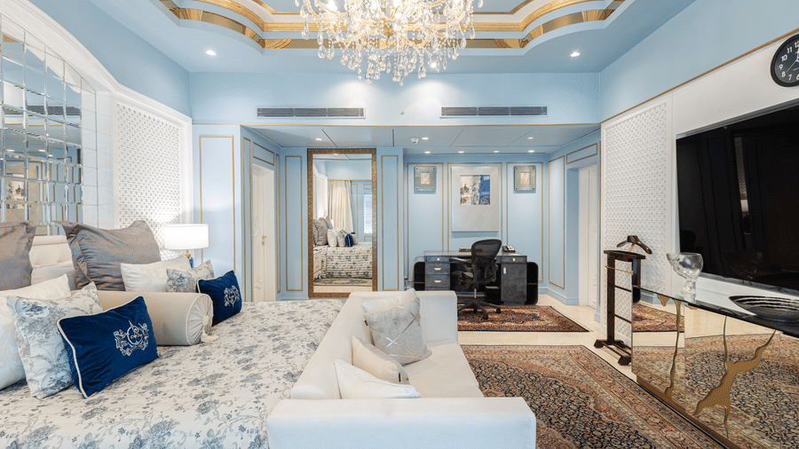 an image of our diplomatic suite room with a large bed with pillows, a couch in front of a television under a chandelier - The Claridges New Delhi
