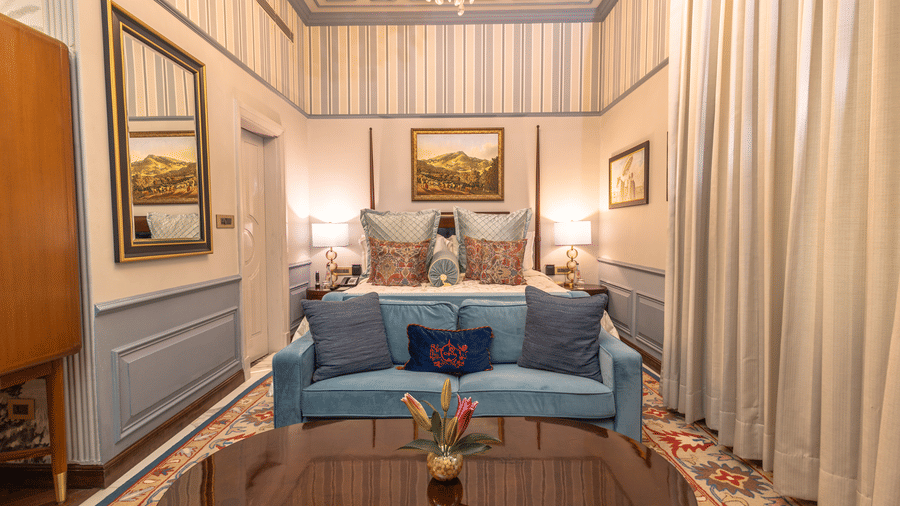 image of the bedroom of the heritage parlour suite with a king sized bed and a blue couch with two cushions placed on it - The Claridges New Delhi