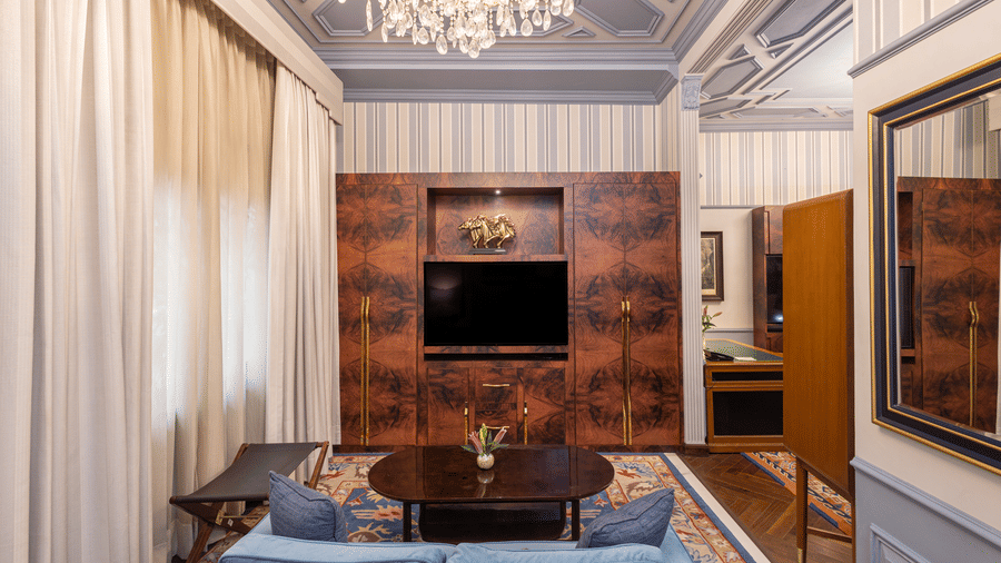 image of the bedroom with a wall mounted television placed in front of the bed with a light blue coloured couch at our heritage parlour suite - The Claridges New Delhi