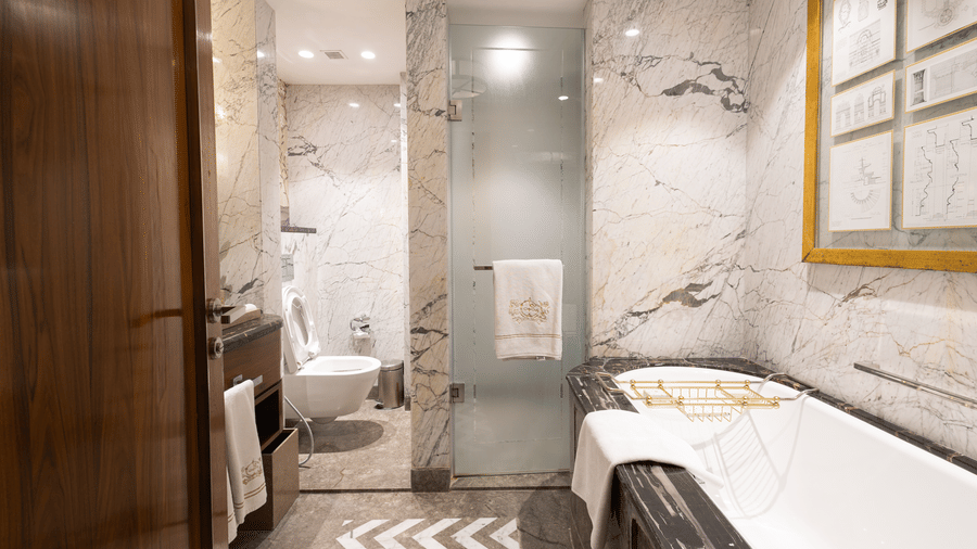 image of the bathroom of the cabana room with white gradient marbles featuring a full length bath tub and a cubical shower  - Image of the Cabana Room featuring two single-seater couches and a wall-mounted television - The Claridges New Delhi