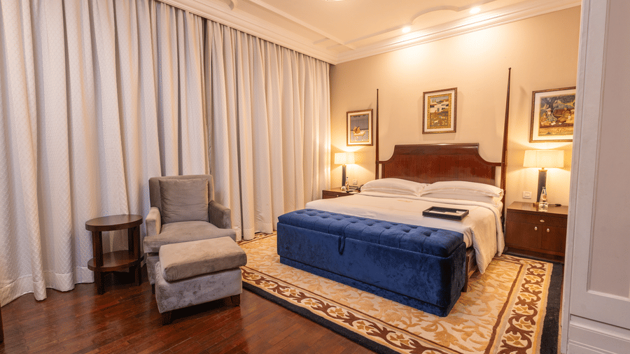 corner view of the luxurious bedroom at our heritage rooms with white curtains and a grey arm chair  - The Claridges New Delhi