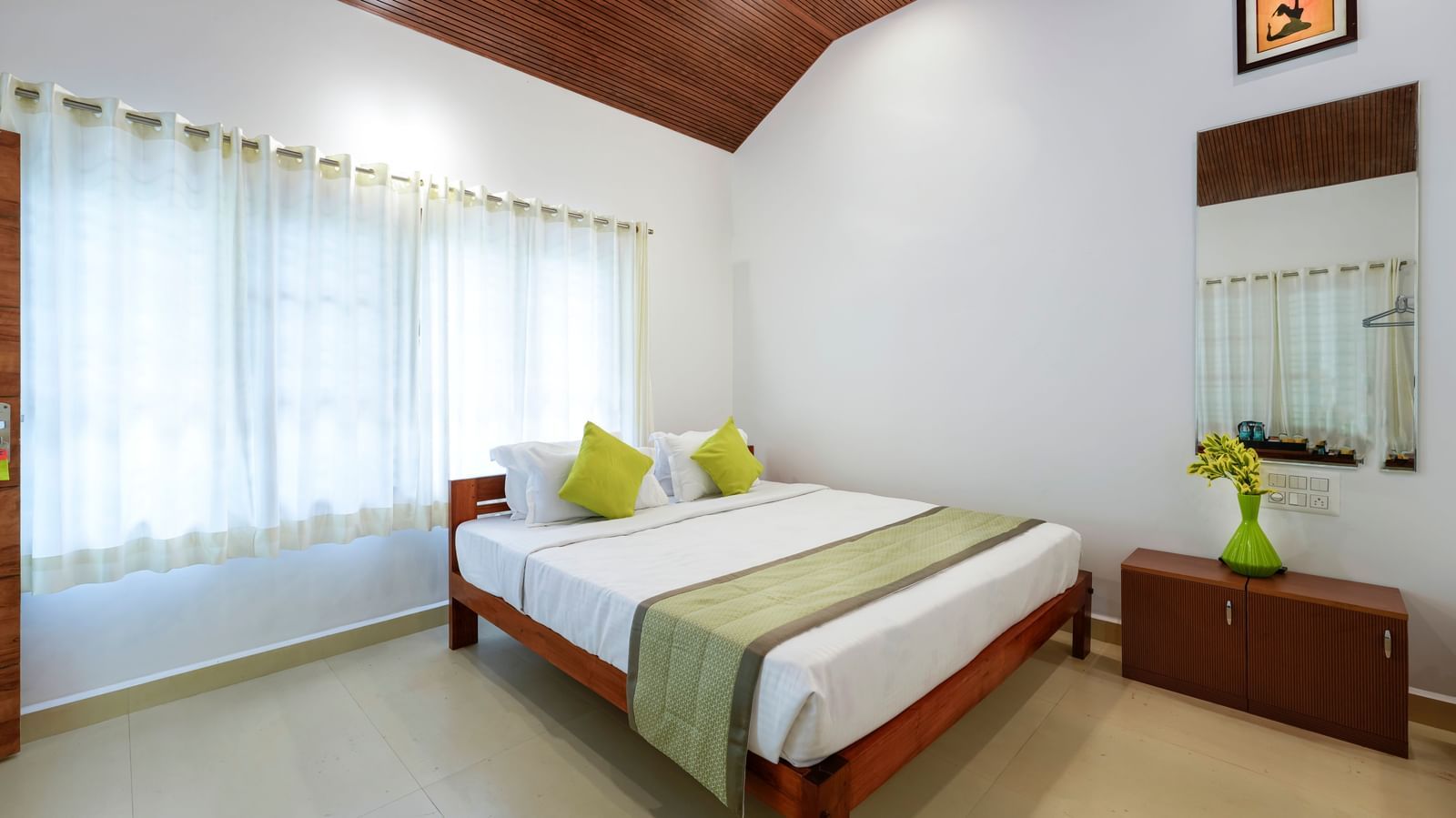 A spacious family room at Bhuthanakadu Retreat, highlighting a double bed and a single bed with bright pillows, large windows with sheer curtains, and a high-pitched wooden ceiling.