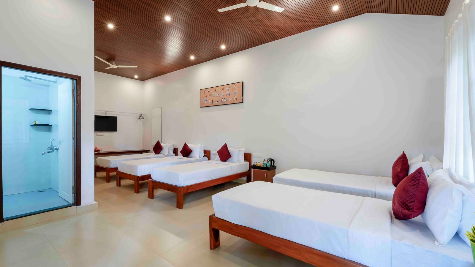 A spacious hall of residences at Bhuthanakadu Retreat, showcasing four single beds with white bedding and red pillows, a modern bathroom, and a warm wooden-paneled ceiling.