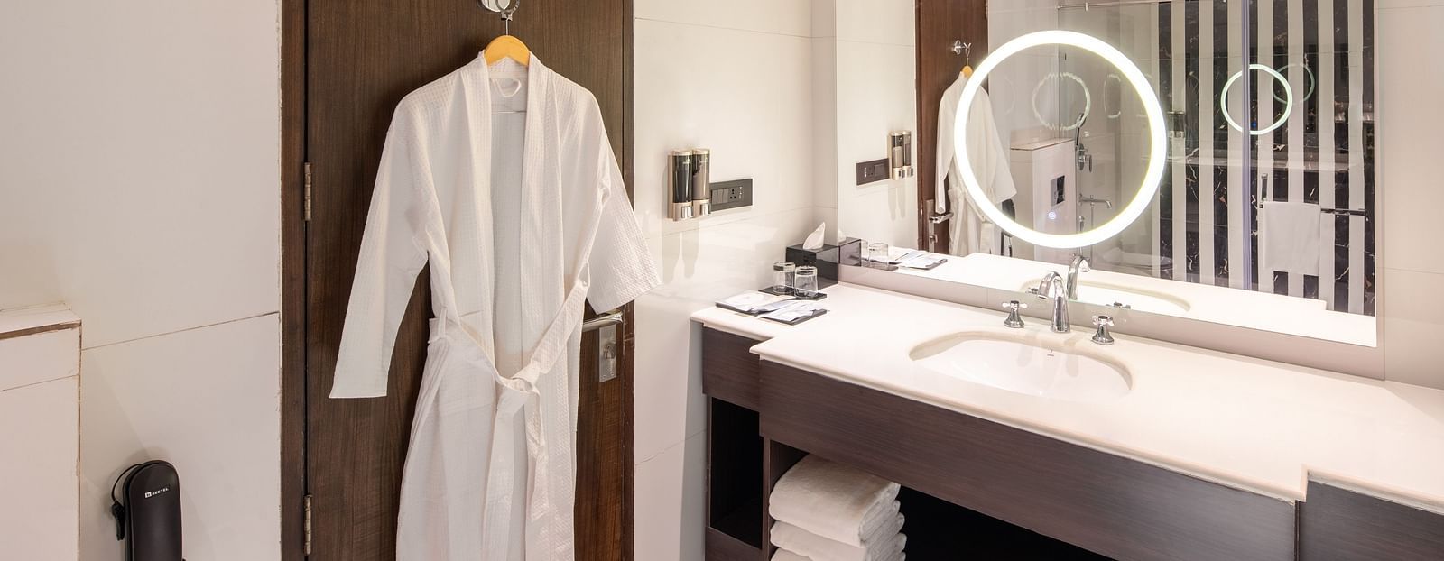 An image of a bathroom with bathrobe hanged on a door, mirror on the wall, cabinet filled with towels and toiletries, and on top of the cabinet there is a ring light - Brij Eternity, Vrindavan, 
