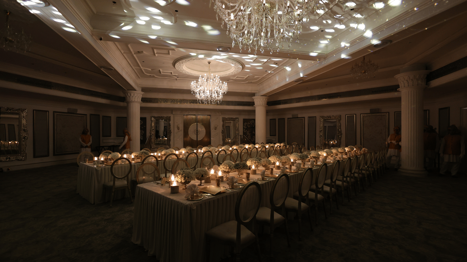 Dimly lit luxury banquet hall featuring a long dining table setup with candles and decorative ceiling light projections under chandeliers.
